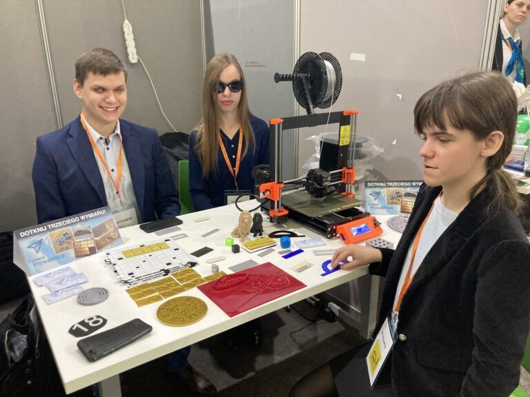 Pierwszy dzień Targów Dostępności. Na stoisku stoi drukarka, leżą także różne przykładowe modele. Przy stoisku stoją Dawid, Maja oraz Monika.