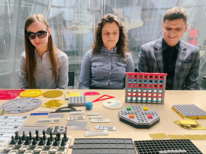 Stoisko z perspektywy zwiedzającego. Monika, Dawid i Juita siedzą przy stole z wydrukami.