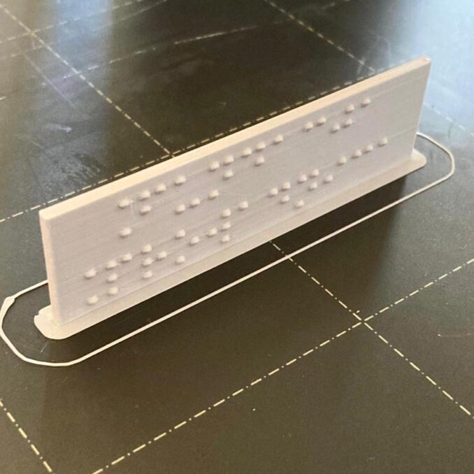 Tabliczka z brajlowskim napisem Fundacja Prowadnica stoi na drukarce 3d, właśnie skończył się wydruk