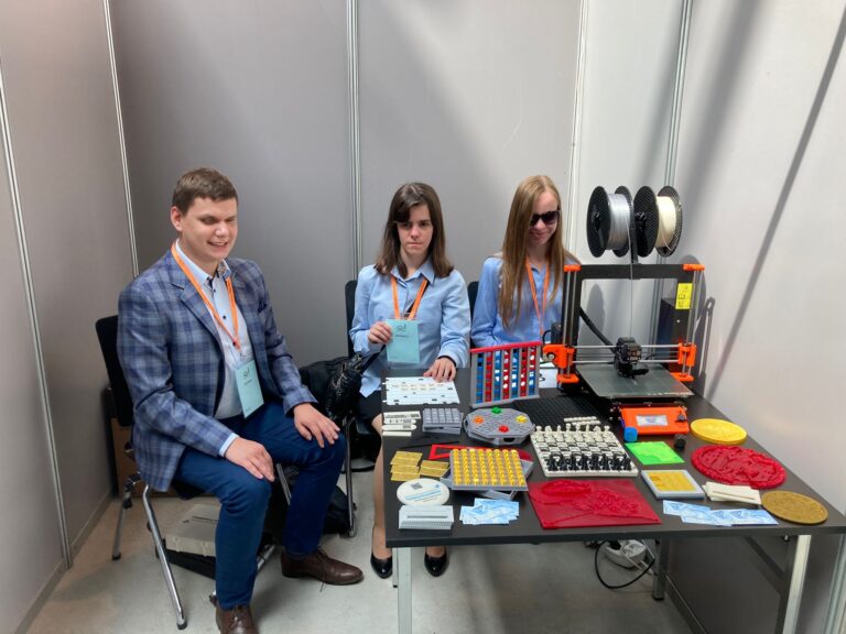 Dawid, Maja i Monika przy stanowisku z dalszej perspektywy. Widać cały stół i drukarkę. Maja trzyma w ręce plakietkę wystawca przodem do widza.