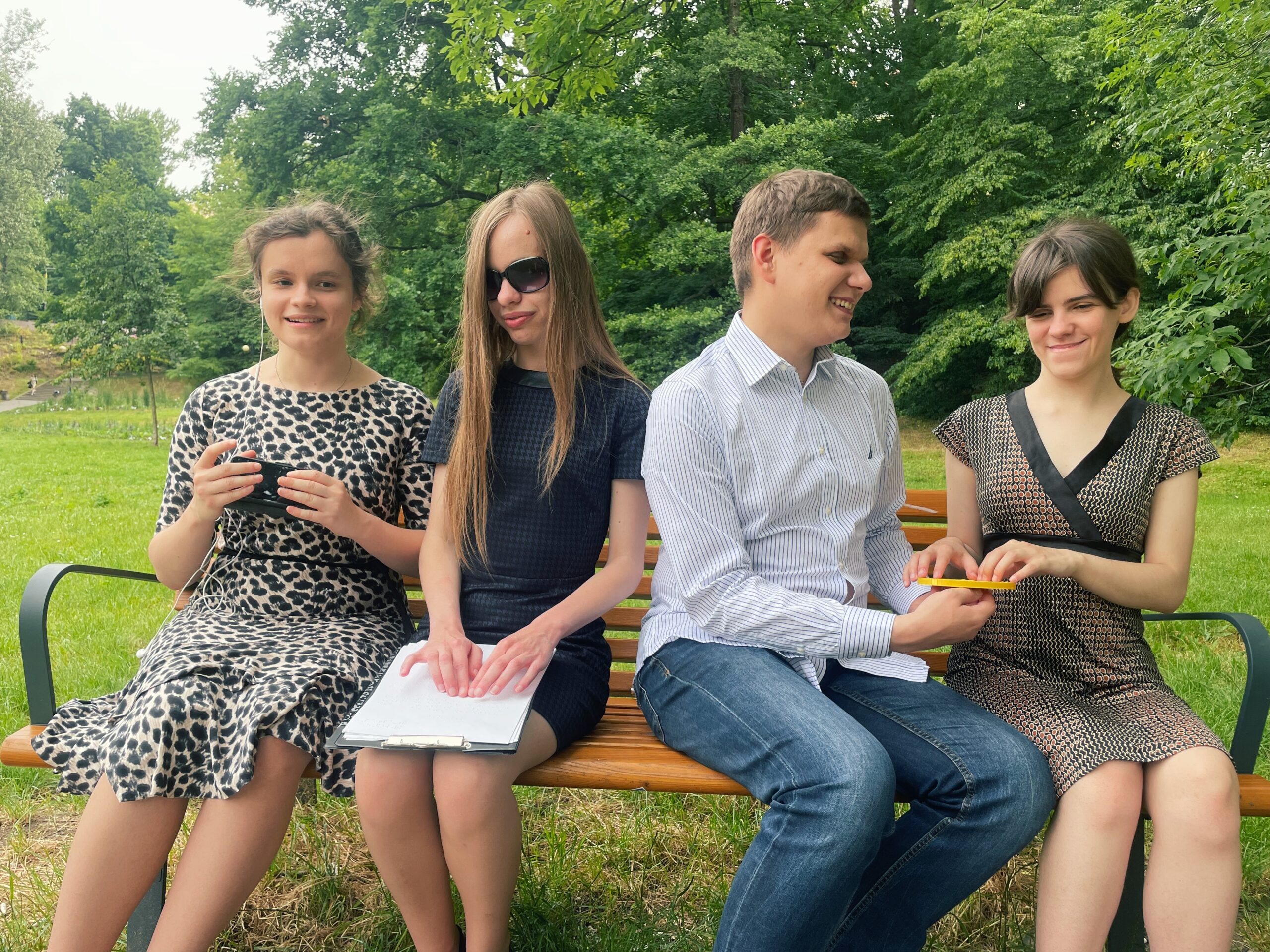 Zarząd siedzi na ławce w parku. Julita pisze coś na telefonie, Monika czyta brajlowski tekst na kartce papieru, Dawid pokazuje Mai jeden z wydruków.