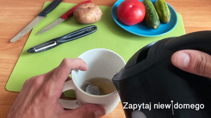 Widok stołu z zieloną podkładką. Na pierwszym planie dłonie, nasz grafik przyznał, że jego własne, ;) wlewają wrzątek do kubka: prawa trzyma czajnik, palec wskazujący lewej oparty jest o wewnętrzną stronę kubka. Dalej, po lewej, leżą ostrzami do widza dwa noże: jeden domasła, drugi ostrzejszy i obieraczka. Obok obieraczki nieobrany ziemniak. Po prawej stoi talerz z pomidorem i dwoma ogórkami.