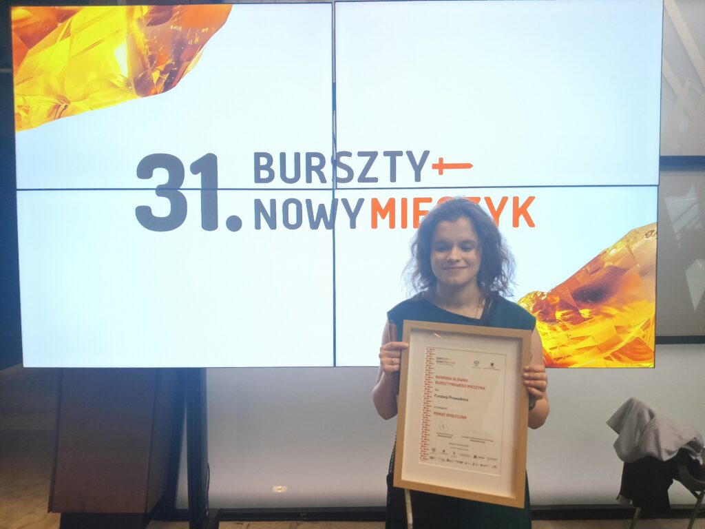 Julita, w wieczorowej sukni, stojąca na tle tablicy multimedialnej z napisem 31 bursztynowy mieczyk, z nagrodą w rękach