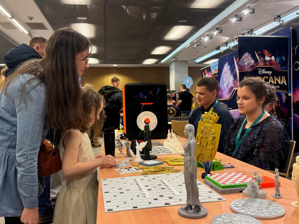 Rodzice z dziećmi oglądają druki i rozmawiają z zarządem. Widać stoisko, drukarkę, banery, ogrom ludzi i wystawców