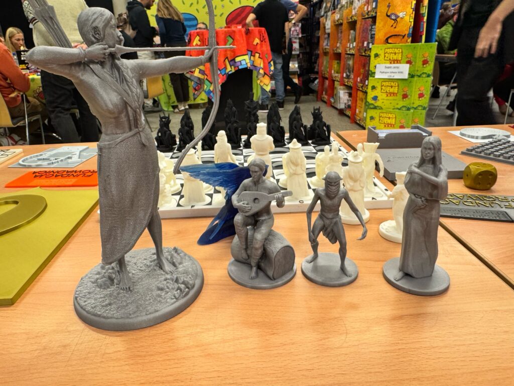 Na stoisku znajdują się wydrukowane z żywicy figurki postaci fantasy: elfka, krasnolud, dziewczyna z łukiem na gałęzi drzewa, grajek z lutnią, ork