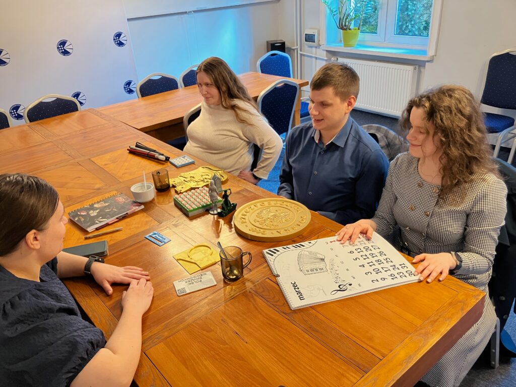 Dawid, Julita i dwie kobiety siedzą przy stole w sali z logami PZN na ścianach. Na stole leżą nasze wydruki, biała laska i kubki. Julita ogląda wypukły kalendarz.