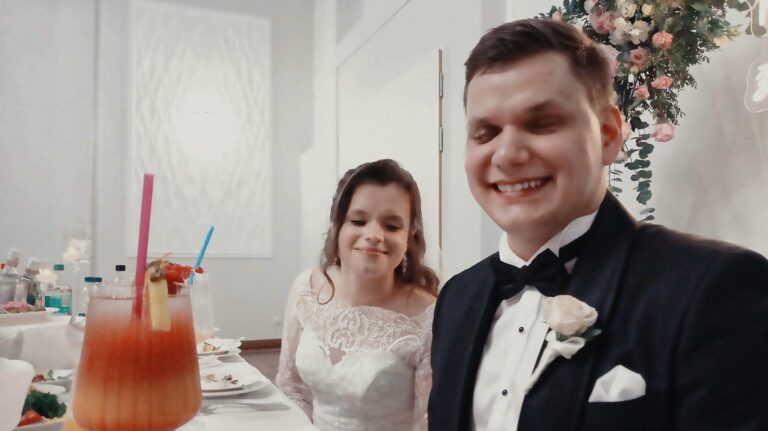 Dawid i Julita siedzą obok siebie przy weselnym stole prezydialnym. Dawid, w czarnym smokingu z muchą i jasnoróżową różą w butonierce, szeroko się uśmiecha, przymykając oczy. Julita, w białej koronkowej sukni ślubnej z długimi rękawami, z upiętymi w stylu romantycznym włosami, delikatnie się uśmiecha. Przed nimi na białym obrusie stoją talerze po posiłku i wysoki kieliszek z kolorowym, warstwowym drinkiem z kawałkiem ananasa i słomką. W tle widać jasną ścianę oraz dekorację z zielonych liści i różowo-kremowych kwiatów, tworzących elegancką oprawę ich stołu.
