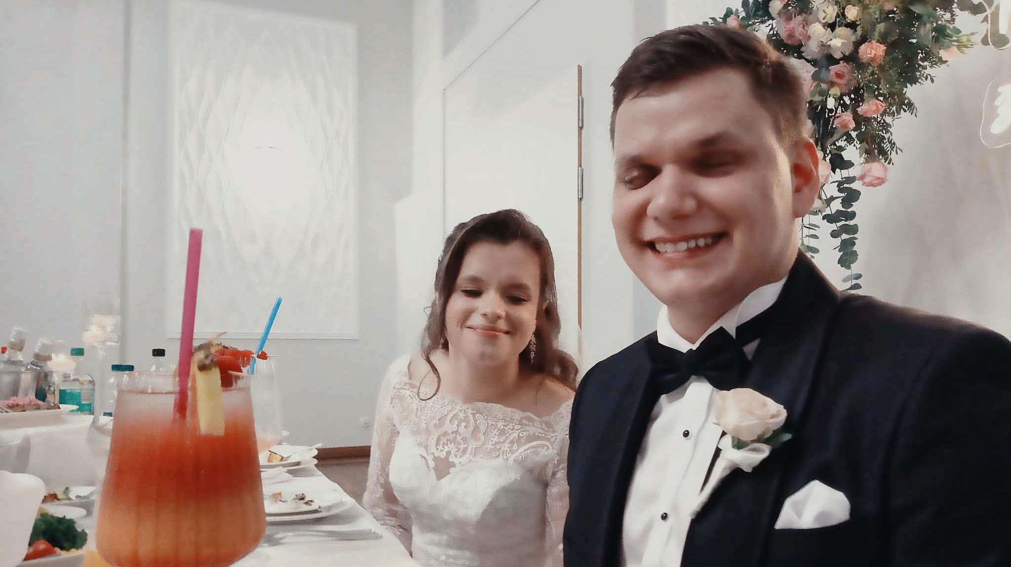 Dawid i Julita siedzą obok siebie przy weselnym stole prezydialnym. Dawid, w czarnym smokingu z muchą i jasnoróżową różą w butonierce, szeroko się uśmiecha, przymykając oczy. Julita, w białej koronkowej sukni ślubnej z długimi rękawami, z upiętymi w stylu romantycznym włosami, delikatnie się uśmiecha. Przed nimi na białym obrusie stoją talerze po posiłku i wysoki kieliszek z kolorowym, warstwowym drinkiem z kawałkiem ananasa i słomką. W tle widać jasną ścianę oraz dekorację z zielonych liści i różowo-kremowych kwiatów, tworzących elegancką oprawę ich stołu.
