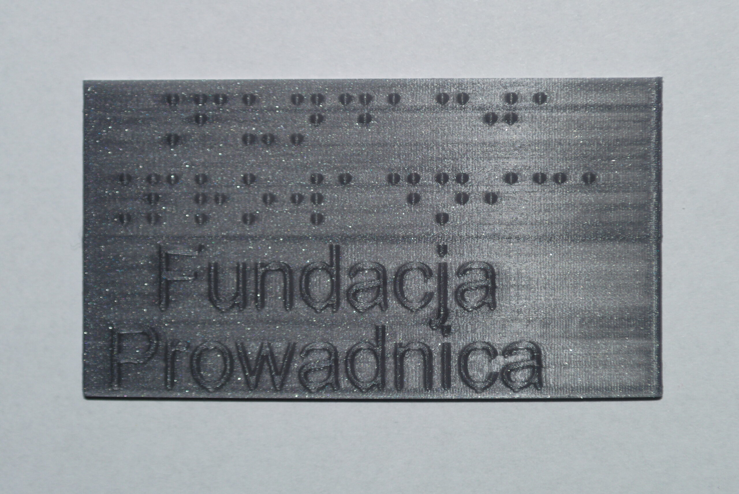 Tabliczka z szarego PLA, z brajlowskim i czarnodrukowym napisem "Fundacja Prowadnica"