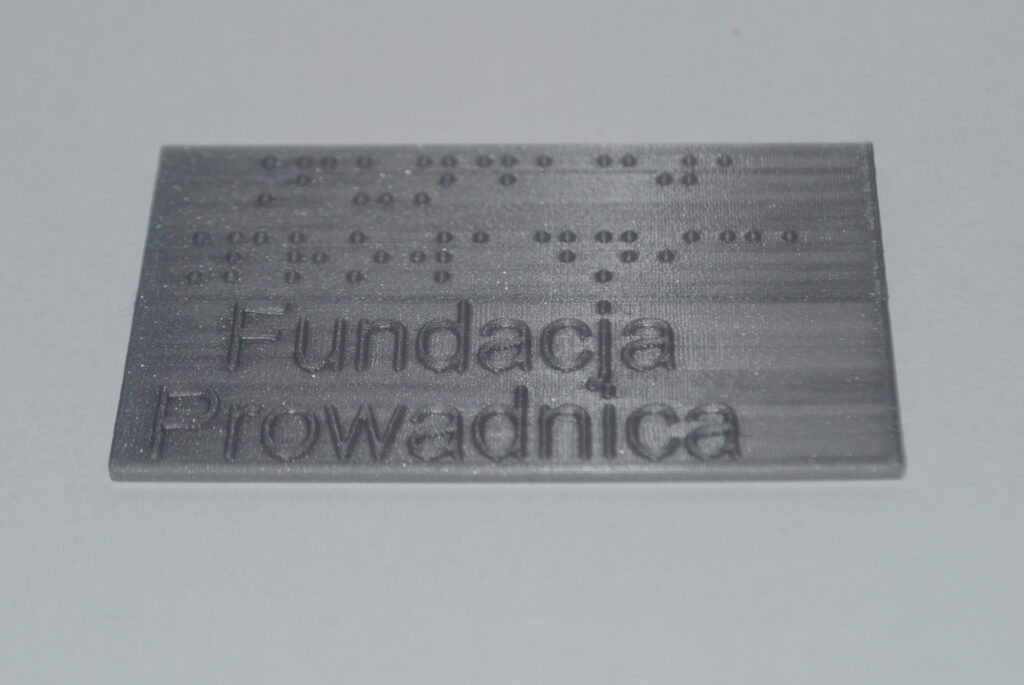 Tabliczka z szarego PLA, z brajlowskim i czarnodrukowym napisem "Fundacja Prowadnica", ujęcie pod kątem
