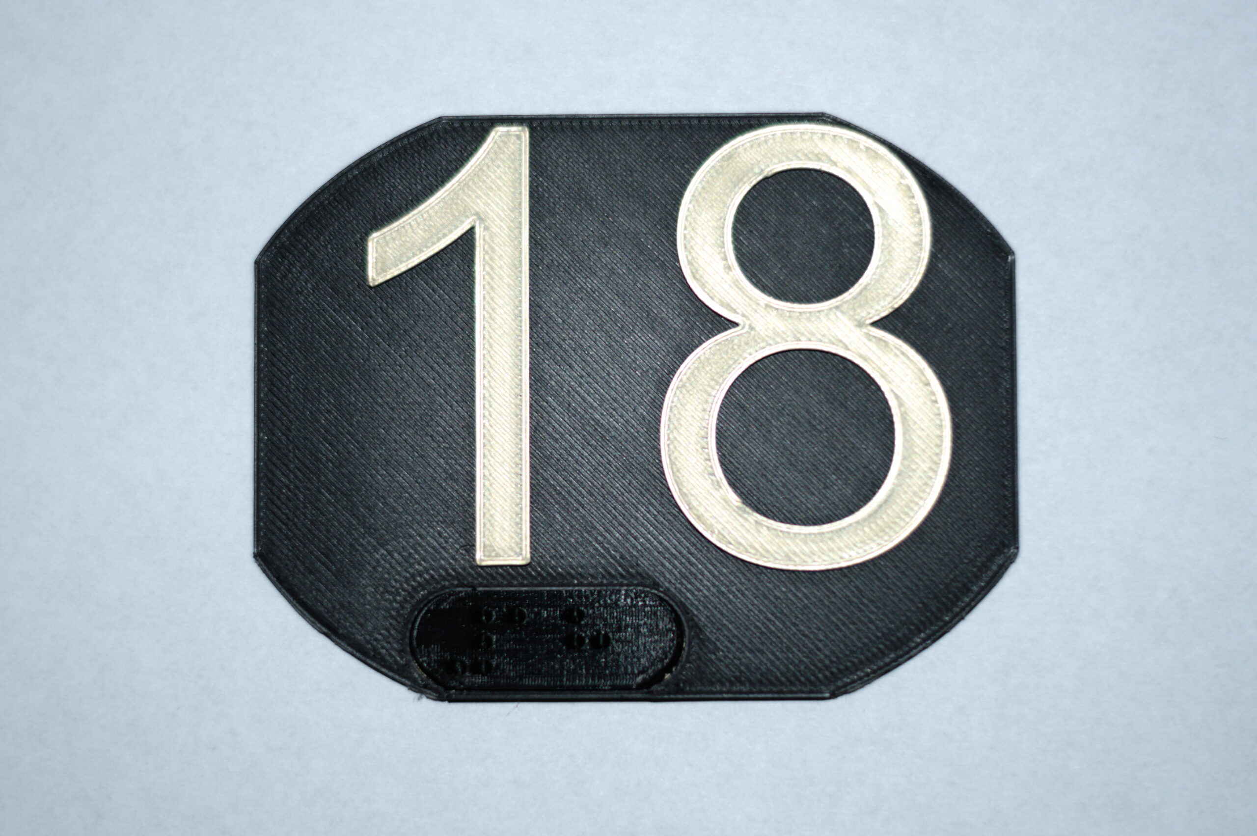 PLA, czarna, duża, zaokrąglona tabliczka z białą liczbą 18 i czarnym napisem w Brajlu