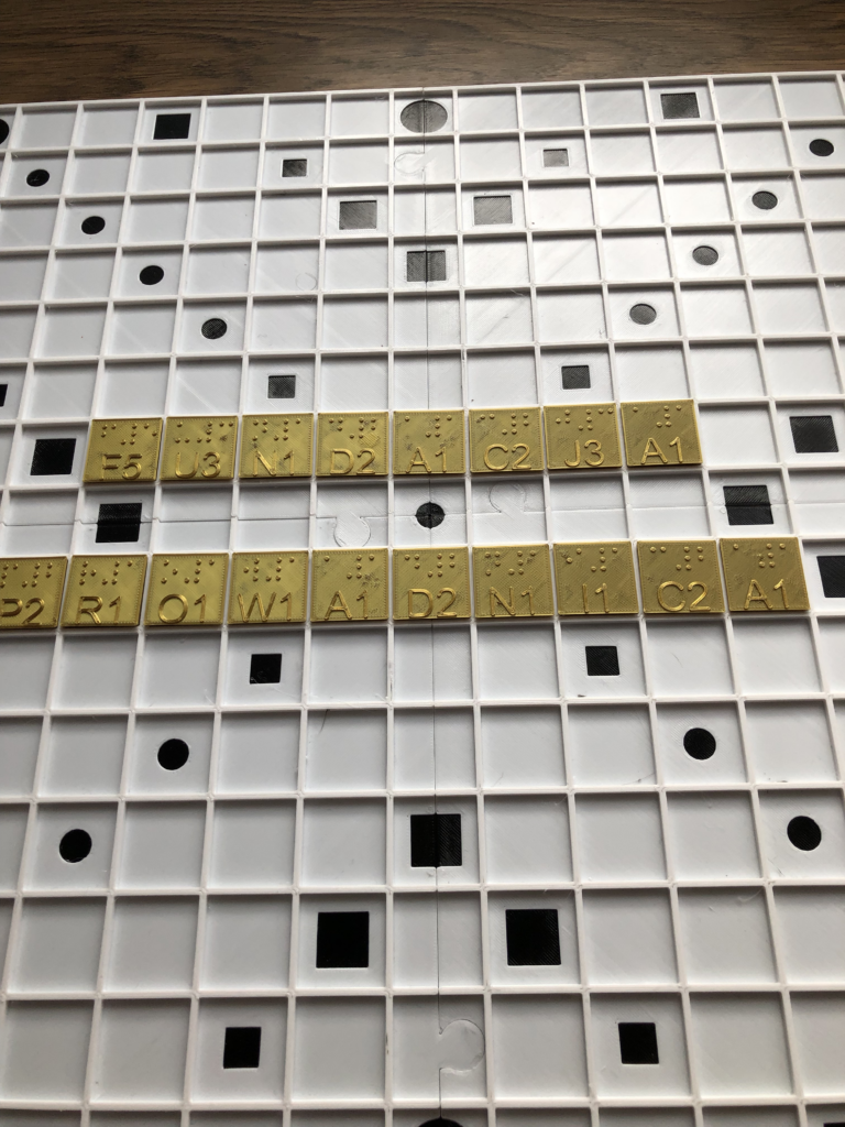 Na planszy do Scrabbli złote litery tworzą napis Fundacja Prowadnica podzielony na dwa rzędy. Litery zawierają brajlowskie oraz czarnodrukowe znak i liczbę punktów.