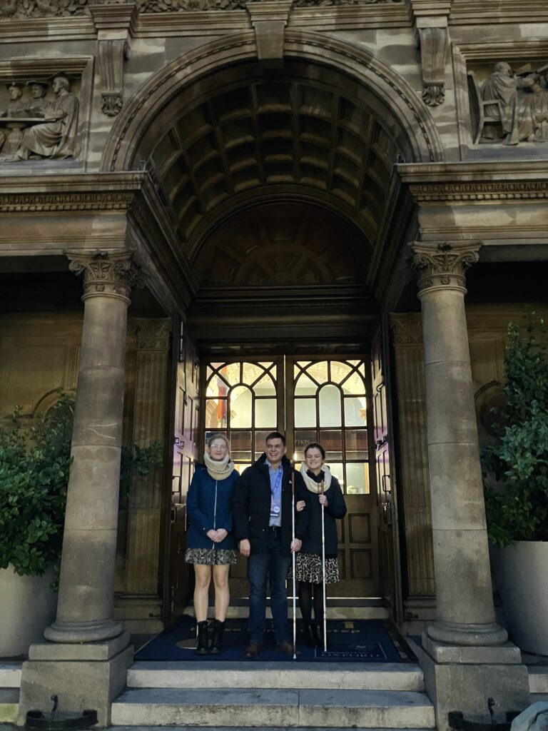 Uśmiechnięci Dawid, Julita i Magda stoją na progu zabytkowego budynku. Widać zieleń przy schodach i ornamenty na ścianach