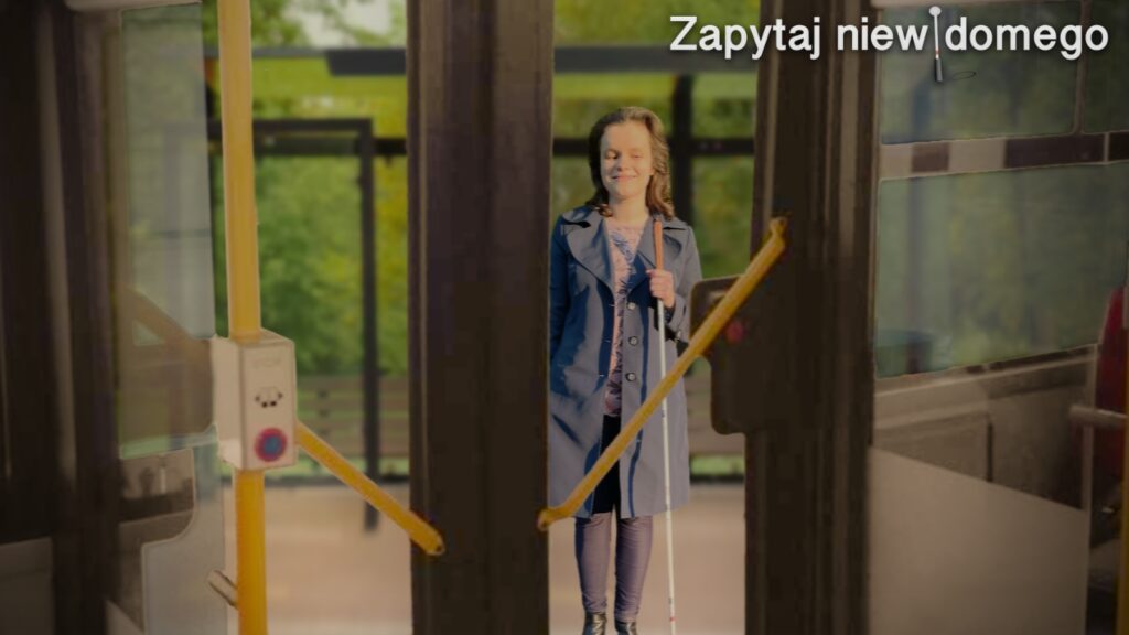 Grafika przedstawia Julitę stojącą na przystanku w wiosenny dzień. Julita ubrana jest w płaszcz, bordowe spodnie i botki. Na zdjęciu widać też drzwi autobusu z charakterystycznymi żółtymi poręczami i przyciskami do żądania przystanków. Julita trzyma w ręce białą laskę i jest gotowa do wejścia do autobusu.