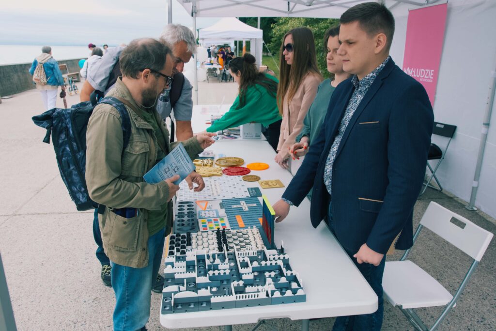 Piknik w Gdyni. Widać stół z wydrukami, stojący za nim zarząd i stojących przed nim dwóch zwiedzających - jeden z których trzyma w ręce ulotkę fundacji. W tle widać wybrzeże.