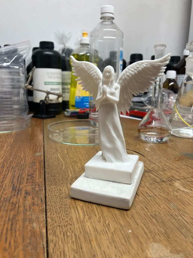 Wygładzona chemicznie figurka białego anioła stoi na drewnianym stole na tle butelek z chemikaliami i sprzętem laboratoryjnym.