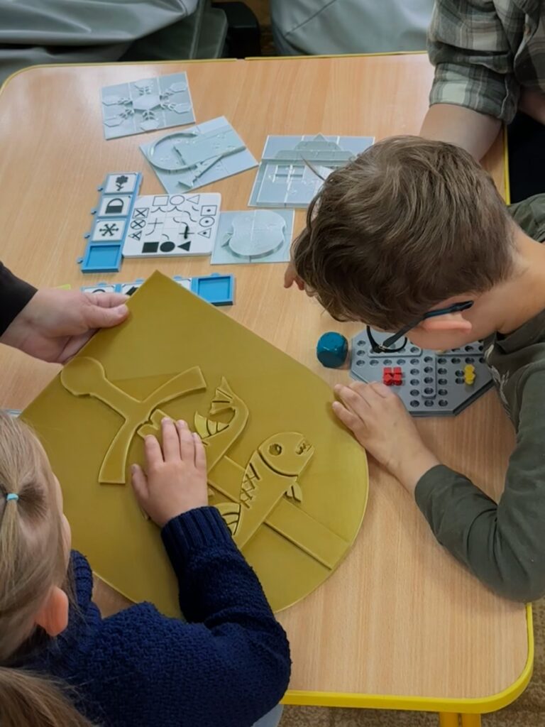 Przedszkole w Laskach: dziewczynka ogląda trzymany przez siostrę zakonną herb Gdyni, a chłopiec z nauczycielką Chińczyka. Na stole widać także puzzle, memory i układankę logiczną „pokaż drogę.”