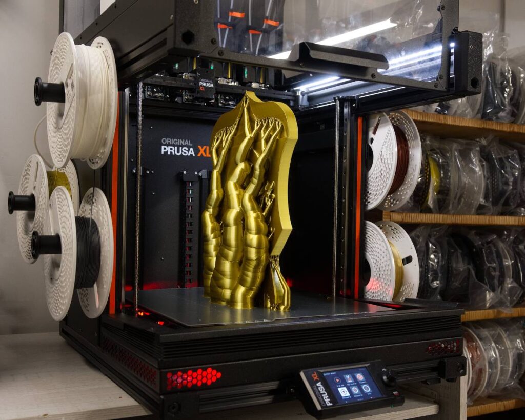 Prusa XL z wydrukowanym z podporami dużym, złotym herbem na stole. Widać, że mimo swojego rozmiaru mógłby być jeszcze wyższy.