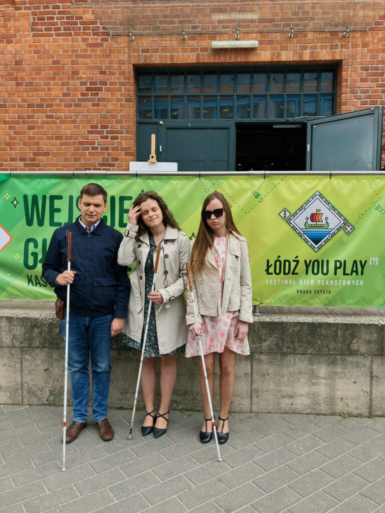 Julita, Monika i Dawid z białymi laskami w rękach stoją na tle ściany budynku z plakatem „Łódź you Play - festiwal gier planszowych”.